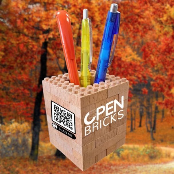 Bunte Stifte in einem Holzhalter mit dem Aufdruck "OPEN BRICKS" vor herbstlicher Kulisse.